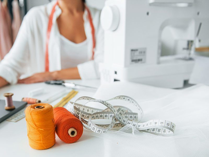 Hilos de colores con una cinta métrica y de fondo una mujer con la máquina de coser